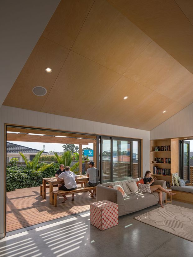 Bright contemporary open-plan living room with a large timber-lined sloping ceiling, polished concrete floor, extensive sliding glass doors, and direct connection to a timber deck. A grey sofa, patterned ottoman, rug, built-in timber shelving, and outdoor dining area are visible, with strong sunlight and garden views.