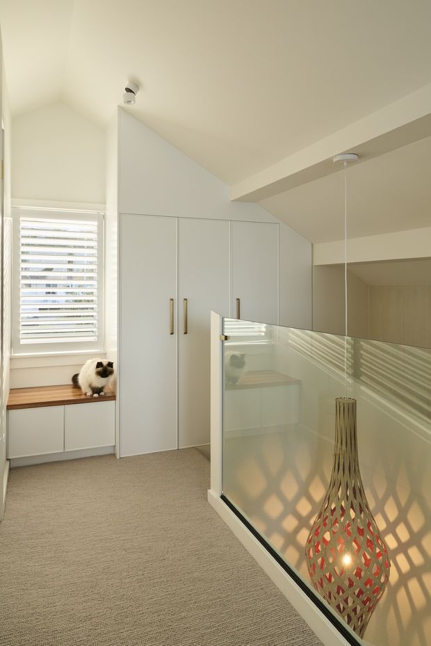 A bright contemporary upper-level hallway or landing with built-in white cabinetry beneath a sloped ceiling, a window seat beside a shuttered window, and beige carpet. A frameless glass balustrade overlooks the lower level, with a decorative pendant light hanging through the void. The space has clean white joinery, soft natural light, and warm neutral finishes.