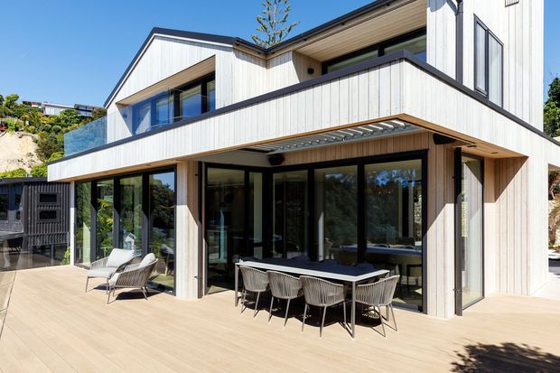 Exterior view of a contemporary multi-level house with pale vertical timber cladding, dark-framed floor-to-ceiling glazing, projecting balconies, and broad roof overhangs. A large timber-toned deck forms an outdoor dining and lounge area with a dining table, chairs, and loungers. The building is set into a sunny landscaped hillside.