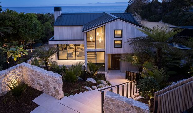 A contemporary multi-level coastal house viewed from an elevated garden path at dusk. The home features vertical timber-like cladding, dark metal pitched roofs, extensive glazed openings, warm interior lighting, landscaped planting, stone retaining walls, and stepped concrete pathways. Ocean and distant islands are visible beyond the property.