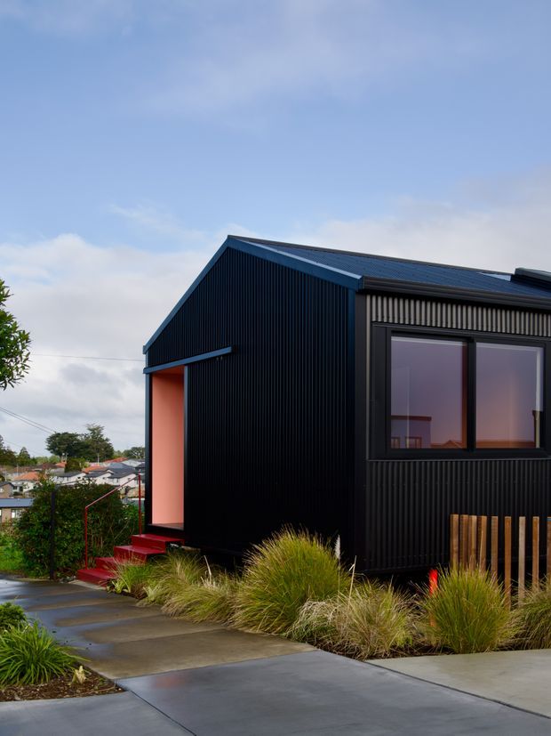 Exterior view of a compact contemporary house with a steep gabled roof and black vertically ribbed cladding. A recessed entry is accented in warm orange-red, with red steps and dark-framed windows. Concrete paving and ornamental grasses lead toward the home, with neighbouring houses visible in the background.