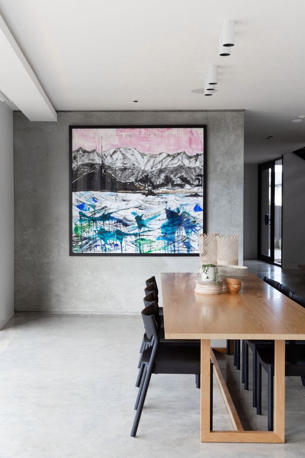 Contemporary dining area with a long light-wood dining table and black dining chairs on a polished concrete floor. A large framed abstract mountain and landscape artwork forms the focal point against a textured grey concrete-look wall. White cylindrical pendant lights hang from the ceiling, with dark-framed glazing and a hallway visible in the background.
