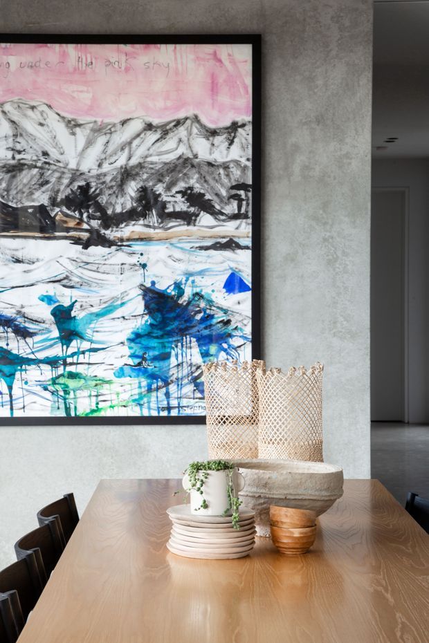 A refined dining area featuring a large timber dining table with dark chairs, layered ceramic tableware, a potted trailing plant, woven decorative vessels, and a substantial stone bowl. The room has a textured grey plaster wall, a large colourful abstract landscape artwork in a black frame, and a glimpse of a neutral hallway beyond.