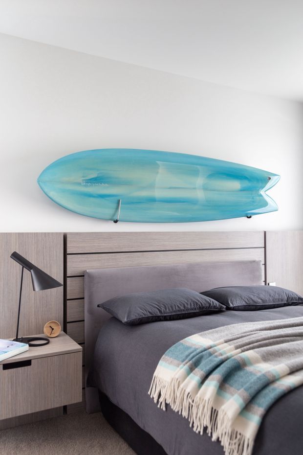 A contemporary bedroom featuring a dark grey upholstered bed with matching pillows, a grey and blue checked throw, and integrated wood-look bedside cabinetry. A large translucent turquoise surfboard is mounted horizontally above the bed as a striking feature. The room has white walls, grey carpet, and a black angled bedside lamp.