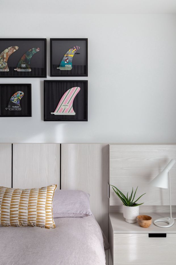 A softly styled bedroom with a pale upholstered bed, light timber wall panelling and integrated bedside cabinetry. Framed surfboard fin artwork decorates the white wall above the bed, while a small potted plant, wooden bowl and white angled lamp sit on the bedside table. The palette is light and restrained with subtle geometric textile accents.