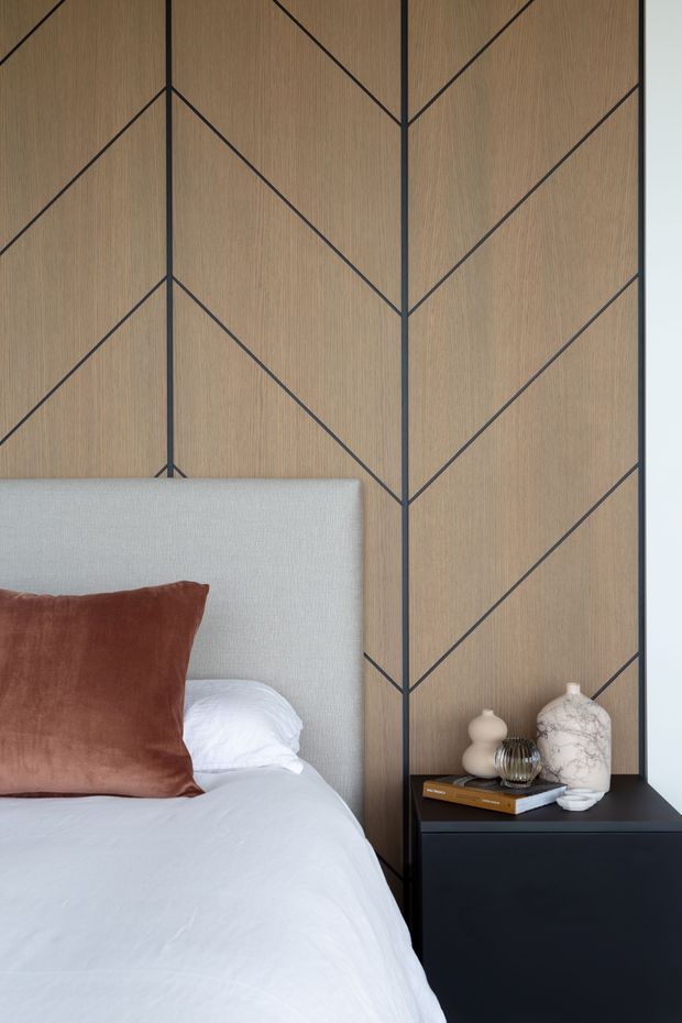 Close-up view of a refined bedroom featuring a light upholstered headboard, white bedding, and a rust-brown accent cushion. The focal point is a full-height warm timber wall panel with dark geometric inlay lines forming a chevron and angular pattern. A dark bedside table holds neutral ceramic and glass decorative vessels and a book. The composition is calm, contemporary, and carefully detailed.
