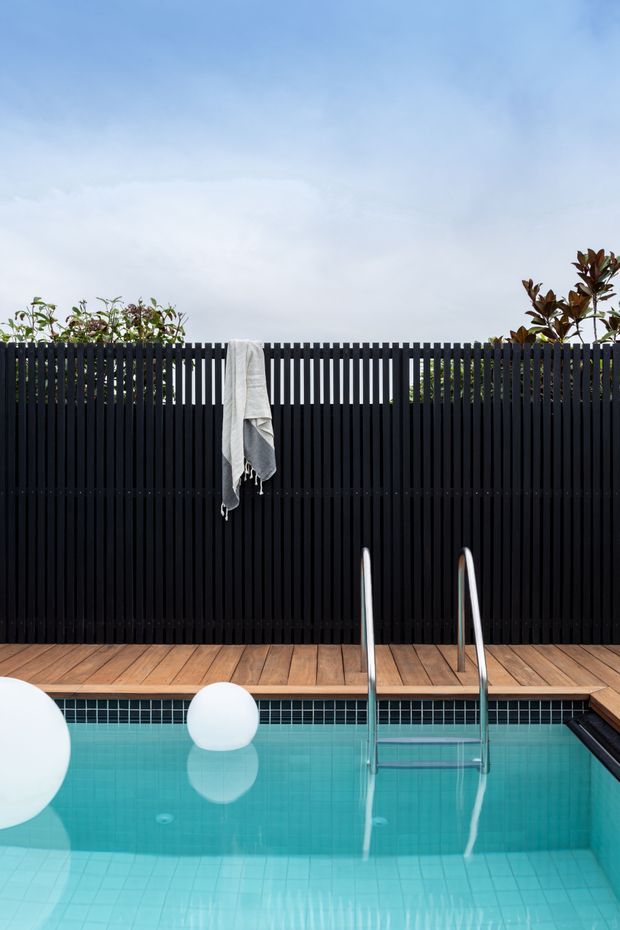 A compact outdoor swimming pool with turquoise tiled water, a stainless-steel ladder, and a timber deck. A tall black vertical-slat privacy fence lines the rear, with planted greenery above it and a towel draped over the fence. White floating pool spheres add a minimalist decorative touch.