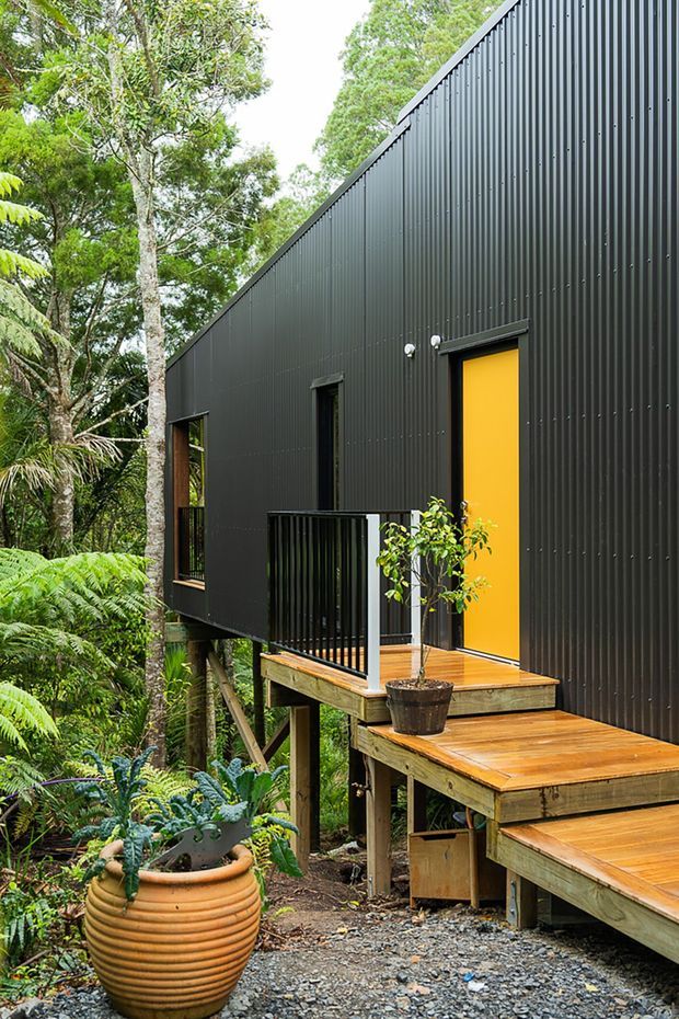 Exterior view of a compact elevated building set among dense native vegetation. The structure has a dark vertically ribbed metal-clad facade, black-framed windows and railings, a vivid yellow entry door, and a stepped timber deck and walkway raised on posts above a gravel and planted garden setting.