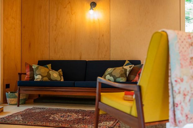 Warm, retro-inspired living room with a dark upholstered sofa against full-height natural timber wall panelling, colourful patterned cushions, a bright yellow mid-century-style lounge chair in the foreground, and a richly patterned area rug. A simple exposed-bulb wall light adds a cozy glow.