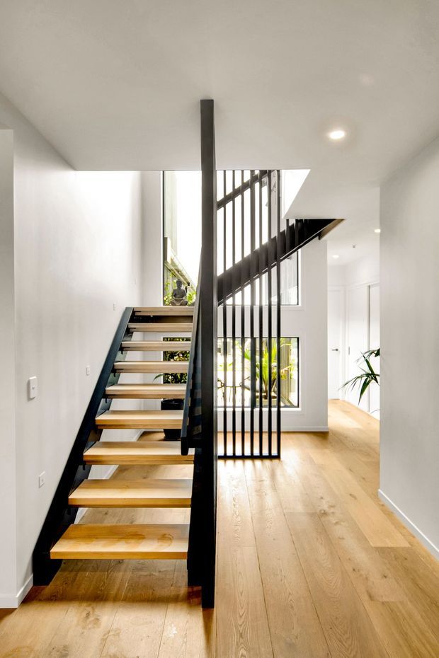 Bright contemporary interior hallway with an open-riser timber staircase, black metal stringers and vertical balustrade, white plaster walls, recessed ceiling lighting, and pale timber flooring. A small courtyard or planted area is visible beyond the stair and glazed openings.
