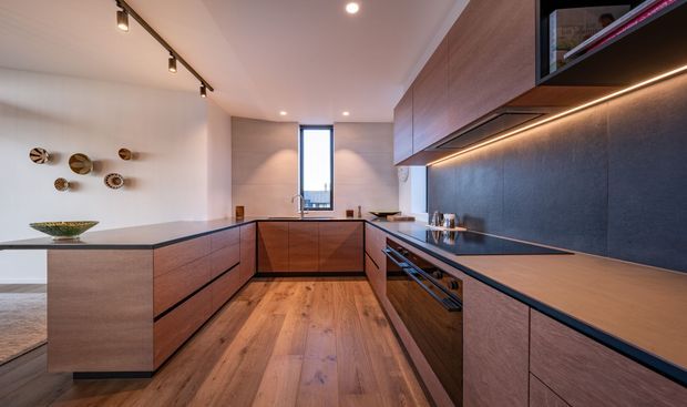 Contemporary galley-style kitchen with warm timber cabinetry, a dark stone-look backsplash and benchtop, integrated appliances, timber flooring, and layered LED and track lighting. A compact island separates the kitchen from the adjacent living area, while a narrow window provides natural light.
