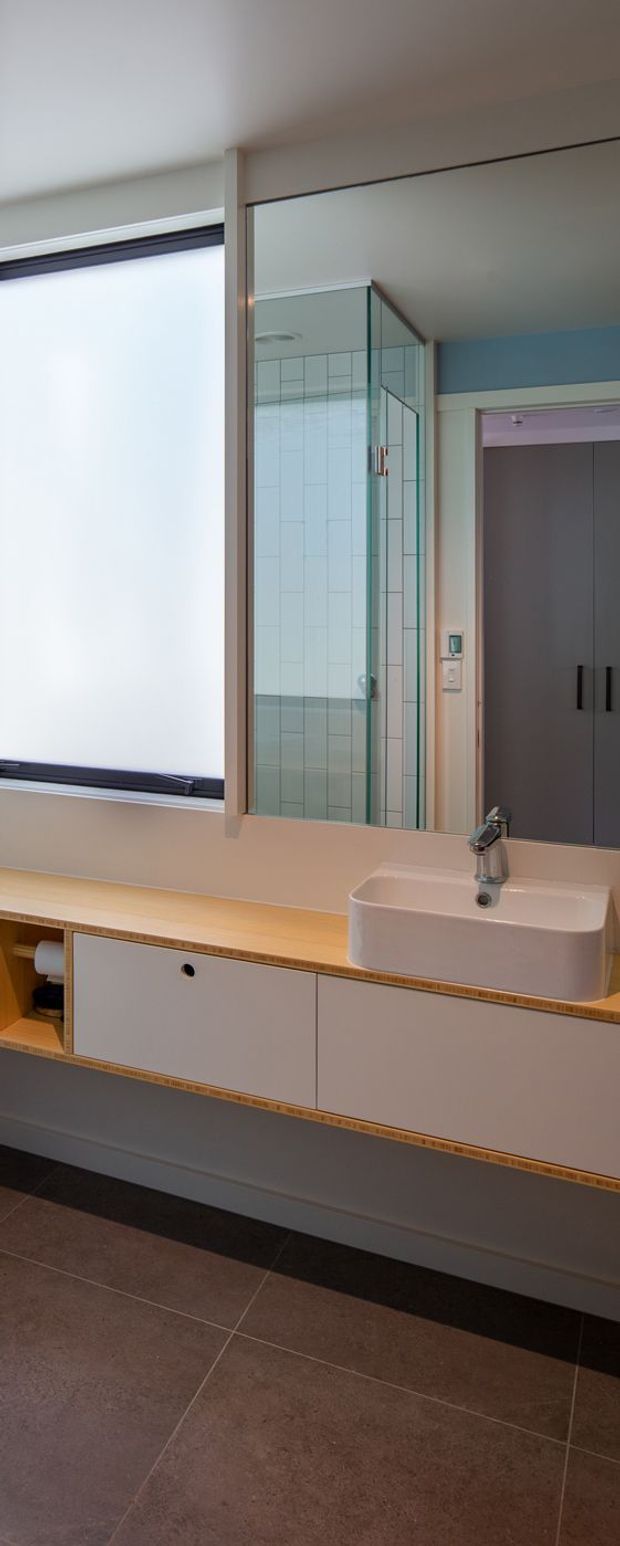 A contemporary bathroom vanity with a wall-mounted, pale grey cabinet featuring integrated timber-edge detailing and a light timber benchtop. A white vessel basin and chrome mixer sit below a large mirror. The mirror reflects a glass-enclosed tiled shower with pale vertical wall tiles. A wide frosted window provides diffuse daylight, and large dark taupe floor tiles ground the room.