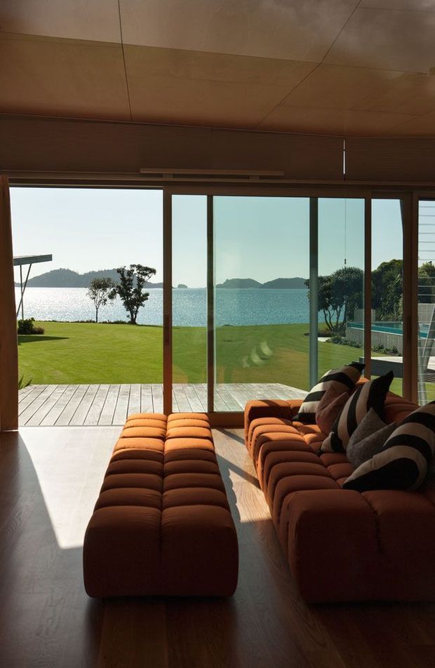 A warm, modern living room opening onto a waterfront lawn and timber deck. The room features a large modular rust-orange sectional sofa with matching ottoman seating, extensive floor-to-ceiling sliding glazing in timber-toned frames, and a wood-finished ceiling and floor. Strong sunlight creates dramatic shadows across the room, while the view includes calm water, distant wooded islands, landscaped grounds, and a swimming pool edge.