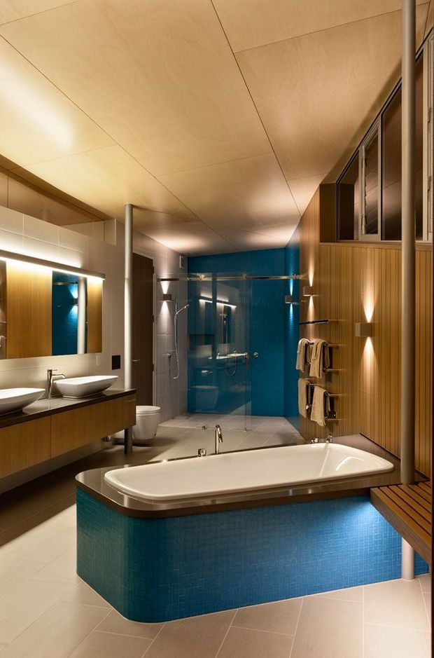 A spacious, warmly lit contemporary bathroom featuring a freestanding rectangular bathtub clad in deep blue mosaic tiles, a glass-enclosed shower, double vessel basins on a timber vanity, large mirrors, timber wall panelling, and integrated ambient lighting. The room has a coordinated palette of blue, warm timber, cream, and soft grey finishes.