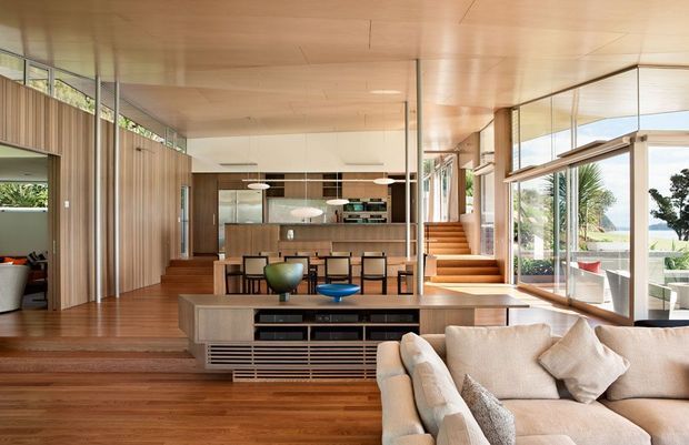 Open-plan modern living and dining area with a warm timber interior. The foreground contains a large upholstered sectional sofa facing a built-in timber media console, while a dining table and kitchen with a central island are visible beyond. Full-height glazed walls and sliding doors open toward a bright landscaped exterior, and timber stairs lead to an upper level. The space has a low, sleek ceiling, extensive built-in joinery, and strong indoor-outdoor connection.