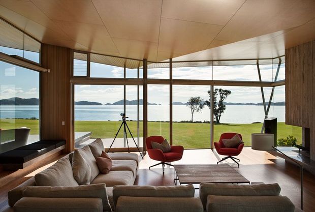 A spacious, light-filled living room with expansive floor-to-ceiling glazing overlooking a calm waterfront landscape. The room features a warm timber-lined ceiling and walls, large timber-framed windows, a broad neutral sectional sofa, red sculptural lounge chairs, and a low timber coffee table. The design has a refined mid-century modern character with strong indoor-outdoor connection and panoramic views.