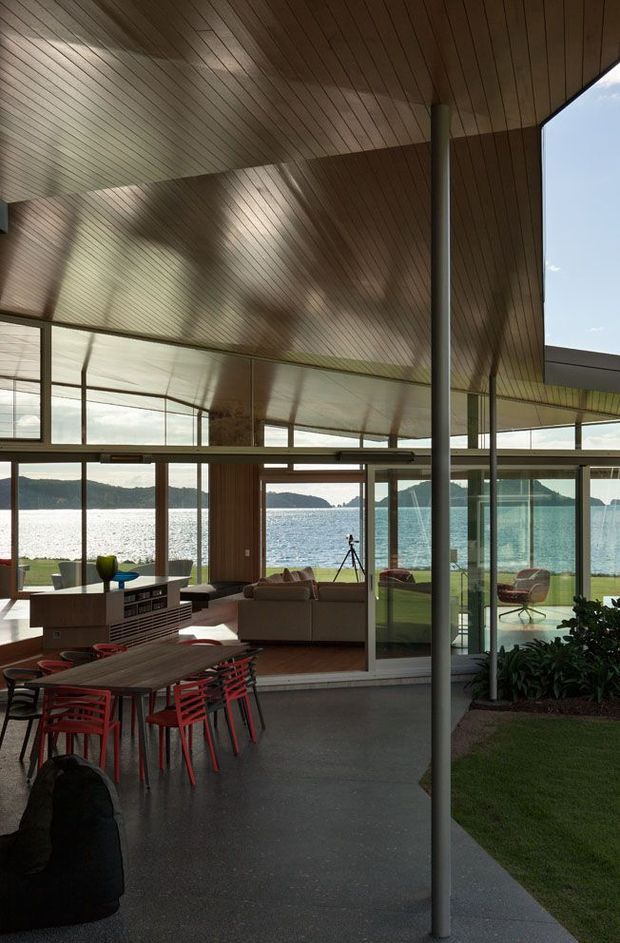 A spacious, open-plan waterfront living and dining area with extensive floor-to-ceiling glazing and sliding glass doors overlooking a bright bay. The room features a dramatic timber-lined ceiling with broad angular planes, slender structural columns, polished concrete flooring, a wood dining table with red chairs, and a built-in island or cabinet separating the dining and lounge zones. A landscaped courtyard edge is visible beside the covered outdoor area.