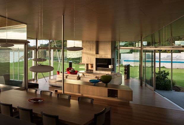 A spacious open-plan living and dining area with extensive floor-to-ceiling glazing overlooking a landscaped garden and swimming pool. The room features a long dark timber dining table, a timber kitchen or media joinery wall, a central fireplace, a low contemporary seating arrangement, pendant lights, and polished timber flooring. The architecture is strongly connected to the outdoors, with slim-framed glazed walls and a low, horizontal modern composition.