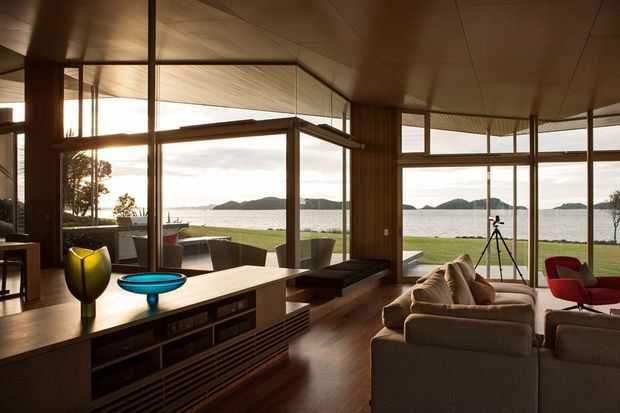 A warm, timber-lined open-plan living room with extensive floor-to-ceiling glazing overlooking a waterfront landscape. The room includes a large sectional sofa, low built-in cabinetry, a central lounge area, and a dining or sitting zone opening onto the lawn. Dark timber floors, timber window frames, and a softly illuminated timber ceiling create a refined mid-century atmosphere, while colourful sculptural decor and a red accent chair add contrast.