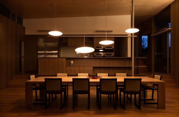 A warm, dimly lit open-plan dining and kitchen interior dominated by a long solid timber dining table with multiple dark upholstered chairs. The room features extensive dark timber wall panelling and cabinetry, a timber ceiling and floor, a linear kitchen with integrated appliances and open shelving, and several suspended white pendant lights. Glazing and a dark exterior view are visible at the right.