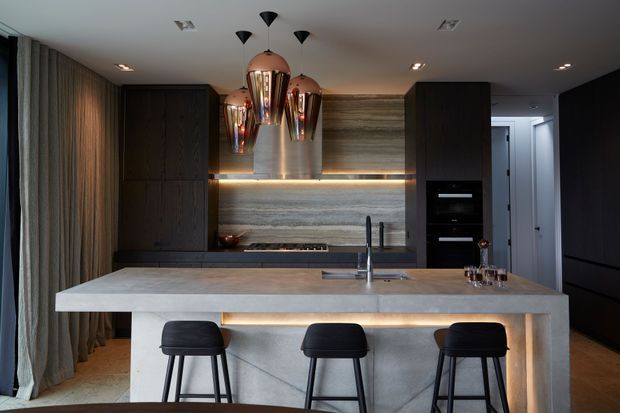 A sophisticated contemporary kitchen with a large pale stone island and three black bar stools. Dark timber cabinetry contrasts with a horizontally textured stone feature wall and illuminated open shelving. Brushed metal pendant lights, integrated appliances, and warm concealed lighting create a refined, moody atmosphere, while full-height curtains line the left side.