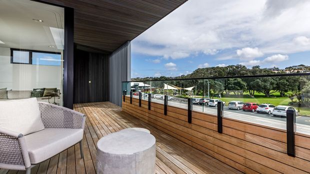 A contemporary balcony or terrace with timber decking and a horizontal timber privacy balustrade topped with clear glass panels and dark posts. The space includes a light upholstered lounge chair and round concrete coffee table beside a fully glazed living area, beneath a timber-lined soffit. A suburban street and landscaped parkland are visible beyond.