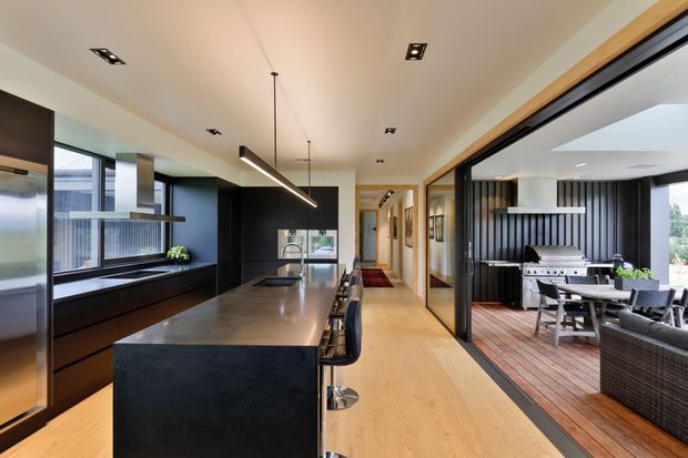 Contemporary open-plan kitchen with a large dark stone island, integrated dark cabinetry, stainless-steel appliances, timber flooring, and linear pendant lighting. Full-height glazing opens to an outdoor dining and barbecue area with dark vertical wall cladding and timber decking.