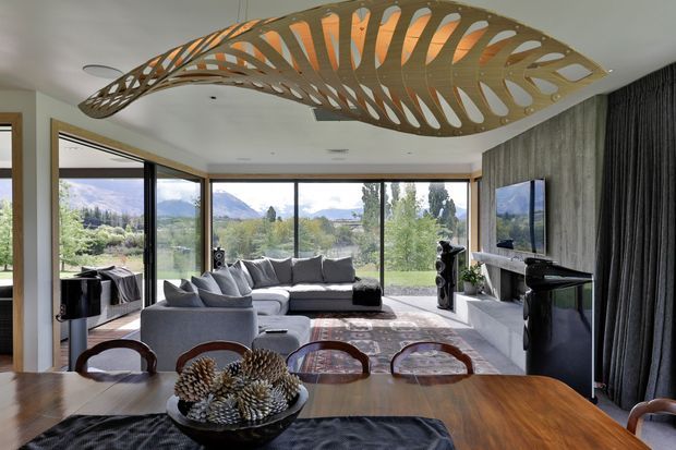 A contemporary open-plan living and dining area with expansive floor-to-ceiling glazing overlooking a landscaped mountain setting. The room features a large grey sectional sofa, media wall with television, dark curtains, timber dining table, black audio equipment, and a striking sculptural timber ceiling installation with an organic curved pattern.