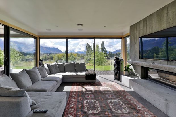 Bright contemporary living room with a large grey sectional sofa, patterned area rug, and sculptural concrete fireplace wall with integrated television. Floor-to-ceiling glazing wraps the room, opening views toward a green landscape and dramatic mountains. The space has a low, horizontal composition and abundant natural light.
