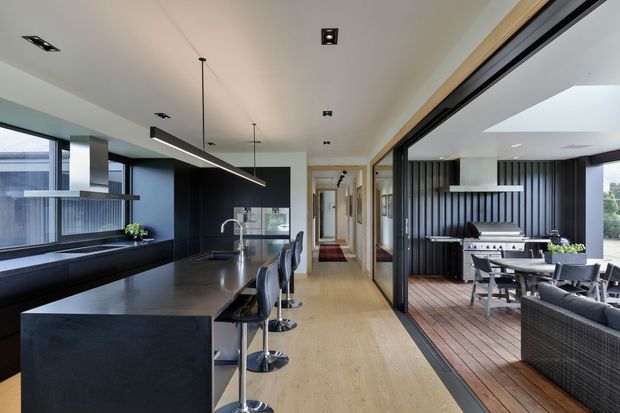 A contemporary open-plan kitchen and living area with a large dark island and integrated breakfast bar, opening through wide glazed doors to a covered outdoor dining and lounge space. The interior features light timber flooring, dark cabinetry, black vertical wall cladding outdoors, large windows, recessed ceiling lights, and a linear pendant above the island.