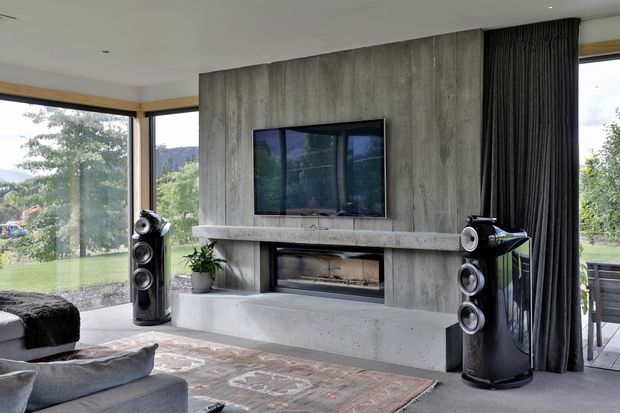 Contemporary living room with a dramatic raw concrete feature wall, integrated linear fireplace, wall-mounted television, large corner glazing, and dark floor-to-ceiling curtains. Sculptural black speakers flank the fireplace, while a sectional sofa, patterned rug, and potted plant create a comfortable media-oriented lounge.
