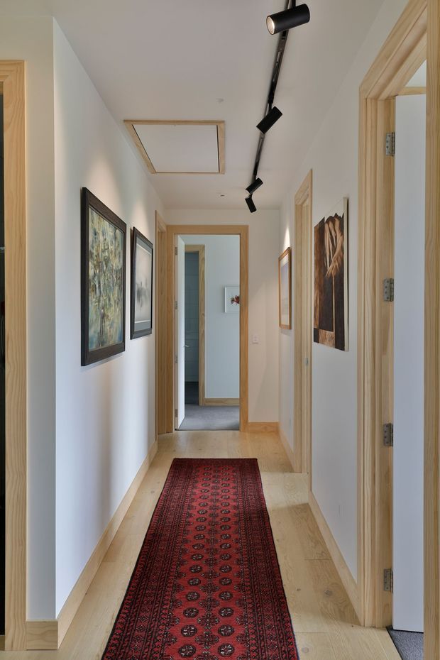 A bright, narrow residential hallway with pale timber flooring and matching timber door frames, white walls, framed artwork, and a long red patterned runner rug. Black track lighting runs along the ceiling, with a recessed skylight or ceiling panel above. Several doorways open into adjacent rooms, including a tiled bathroom or utility space at the end.