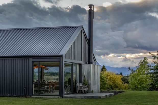 A contemporary single-storey corrugated-metal-clad house with a steep pitched gable roof, large glazed openings, and a prominent black chimney. The building opens onto a timber deck and manicured lawn, with landscaped vegetation and distant mountain views under dramatic clouds.