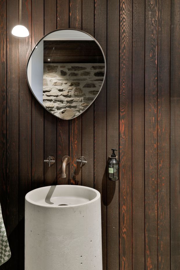 A compact, moody bathroom or powder room featuring a freestanding cylindrical concrete basin, wall-mounted bronze-toned tapware, dark vertically clad timber wall panels, a round mirror, and a small wall light. The mirror reflects a rough stone wall and dark timber ceiling, adding rustic character.