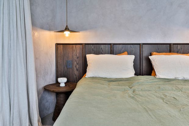 Close-up view of a cozy bedroom with a dark timber-panelled headboard, pale textured plaster wall, soft sage-green bedding, white pillows, and a small dark wood bedside table. A single pendant light hangs beside the bed, with full-height pale curtains at the window. The restrained palette and natural materials create a warm, rustic contemporary atmosphere.