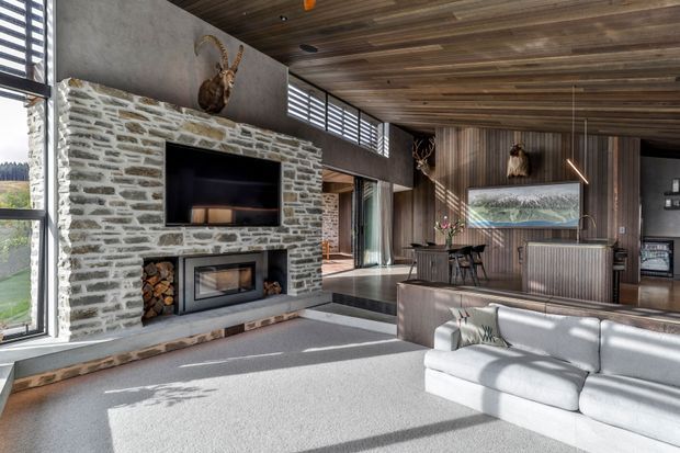 Open-plan contemporary living room with a large white sectional sofa, exposed stone fireplace wall, and television above a built-in wood-burning fireplace. The room includes a timber-clad ceiling and feature wall, high clerestory glazing, a dining area, and a kitchen with a dark island and ribbed metal or timber-fronted cabinetry. Natural daylight creates strong shadows across the pale carpet and polished concrete or stone hearth.