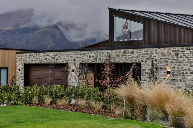 Exterior view of a contemporary mountain home with a low stone-clad frontage, dark vertical timber upper volume, large glazed opening, metal pitched roof, and integrated garage door. Ornamental grasses and structured planting soften the facade, with misty mountains visible behind.