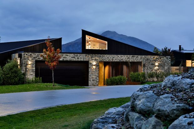 Contemporary single-storey residential facade at dusk, composed of dark gabled volumes, extensive rough natural-stone walling, a recessed dark garage door, and a warm timber-slatted entry or screened living area. Low landscape planting, lawn, driveway, and foreground rockwork frame the house, with a prominent mountainous backdrop visible behind the roofline.