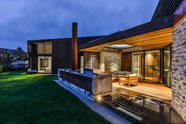 Twilight exterior view of a contemporary single-storey home with dark cladding, extensive glazing, natural stone walls, and a covered outdoor entertaining area. Warm timber soffits and decking surround an outdoor lounge with a built-in fire feature, while landscaped lawn and reflective dark paving lead toward the house.