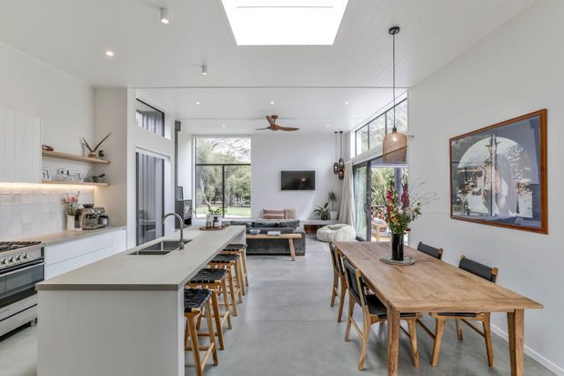 Bright open-plan interior combining a contemporary kitchen, dining area, and living room. A large island with seating faces a spacious dining table, while full-height glazing and a skylight bring in abundant natural light. The room features pale cabinetry, light stone surfaces, polished concrete flooring, timber furniture, pendant lighting, and a neutral palette with indoor plants and artwork.