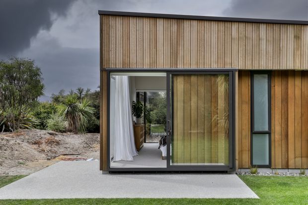 Exterior view of a compact contemporary timber-clad house, featuring vertical natural wood boards, dark metal trim, large sliding glass doors, and a narrow vertical window. The home sits beside a simple concrete path and landscaped garden under an overcast sky.