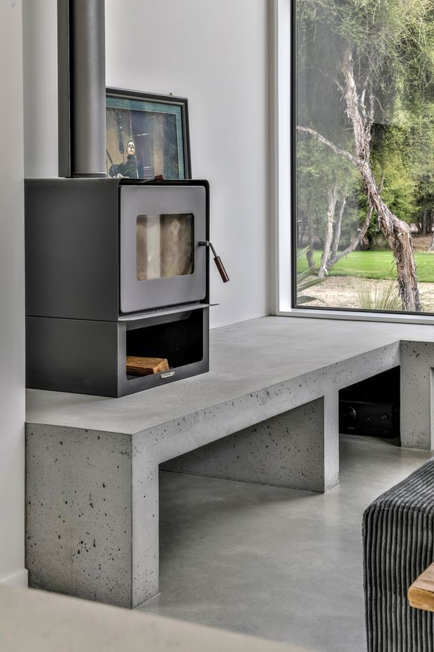 A minimalist living room with a black freestanding wood-burning stove beside a large floor-to-ceiling window. A substantial raw-looking concrete bench or hearth runs beneath the window and stove, with open storage beneath. The room has white walls, a smooth grey concrete floor, dark ribbed upholstered seating at the edge of frame, and views of trees and lawn outside.