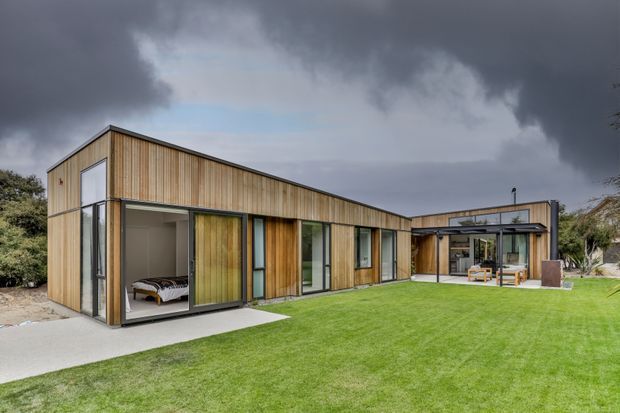 Single-storey contemporary residence with a low, angular gabled roof form and extensive vertical timber cladding. Large glazed openings reveal a bedroom at the left and living or outdoor entertaining spaces at the rear, opening onto a broad manicured lawn. The image is an exterior architectural view under dramatic overcast skies.
