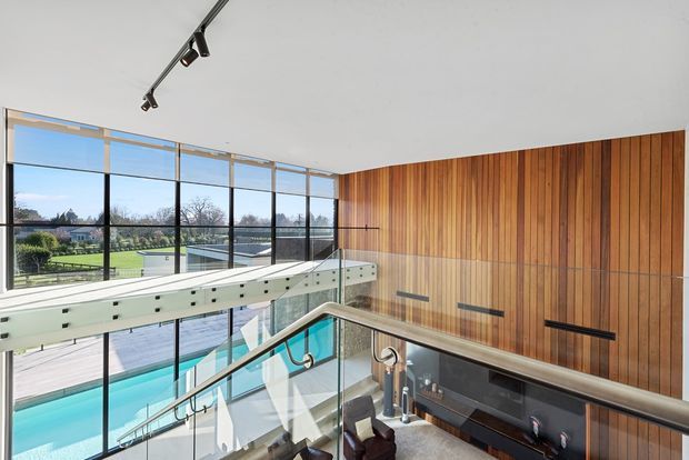 A bright, contemporary multi-level interior overlooking a swimming pool and landscaped grounds. The image focuses on a stairway and upper-level landing with clear glass balustrades, stainless-steel handrails, exposed structural elements, expansive floor-to-ceiling glazing, and a tall vertical timber feature wall. Track lighting runs across the white ceiling.