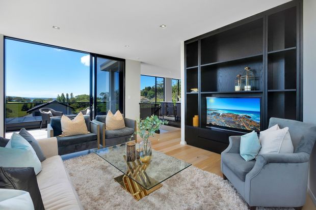Bright contemporary living room with floor-to-ceiling glazing overlooking a green suburban landscape. The room features a large grey sectional sofa, matching armchair, glass coffee table, textured area rug, timber flooring, and a full-height dark built-in media wall with open shelving and television. A balcony or terrace is visible beyond the glazing.