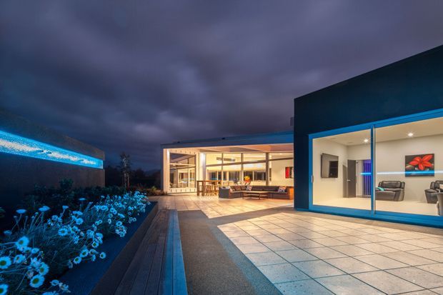 Twilight view of a contemporary home opening onto a broad tiled terrace and landscaped garden. Large blue-framed glazed openings reveal a bright living area, while the recessed central section appears to connect to an open-plan kitchen or dining space. The scene has strong indoor-outdoor integration, low horizontal rooflines, clean geometric volumes, and dramatic blue evening lighting.