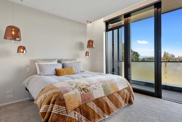 Bright contemporary bedroom with a low-profile bed dressed in white bedding, patterned brown throw, and a mustard accent cushion. The room has pale walls, carpeted flooring, copper pendant lights, and expansive black-framed sliding glass doors opening to a balcony with a distant landscape view.