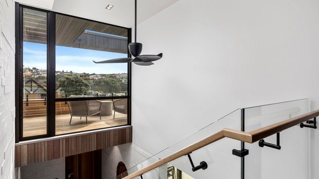 Modern stair landing overlooking a timber-decked balcony and landscaped suburban view through large black-framed glazing. The space features a glass balustrade, timber handrail, black metal supports, a sculptural ceiling fan, white masonry walls, and timber-lined exterior soffit and joinery.