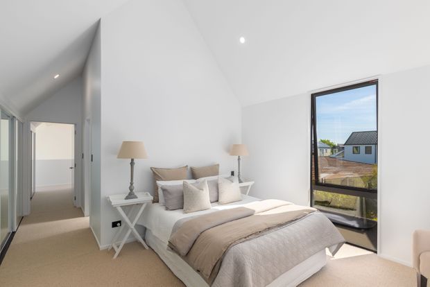 Bright, modern bedroom with a pitched ceiling and clean white walls. A neatly made bed with layered neutral bedding is flanked by white bedside tables and classic lamps. Full-height black-framed glazing opens to an exterior view, while a hallway and doorway are visible to the left. The room has soft beige carpet and a calm, light-filled atmosphere.