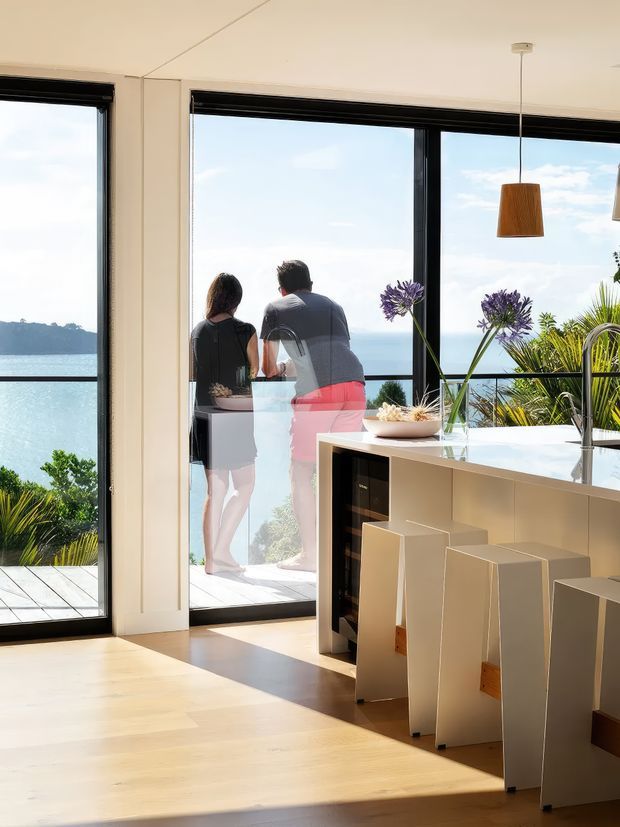 Bright contemporary open-plan kitchen or dining area with a pale timber floor, white cabinetry and island bench, integrated dark appliance joinery, and simple pendant lighting. Full-height black-framed glazing opens to a deck overlooking blue water and a green coastal landscape. Two people are visible on the balcony, and purple flowers and tropical foliage add colour to the interior view.